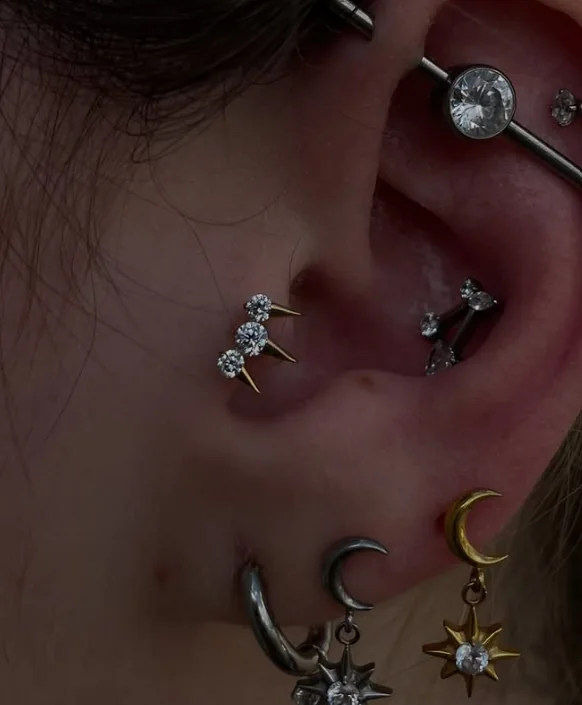 Curadoria de orelha: industrial com zircônia, tragus com trio de pedras e spikes, conch e lóbulos com luas e estrelas