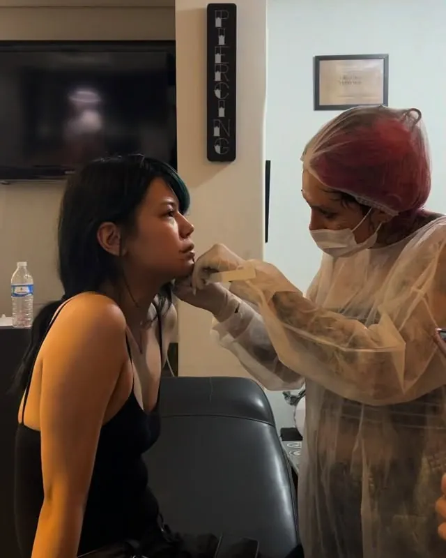 Tali paramentada com touca, máscara e avental fazendo um piercing na orelha de uma cliente