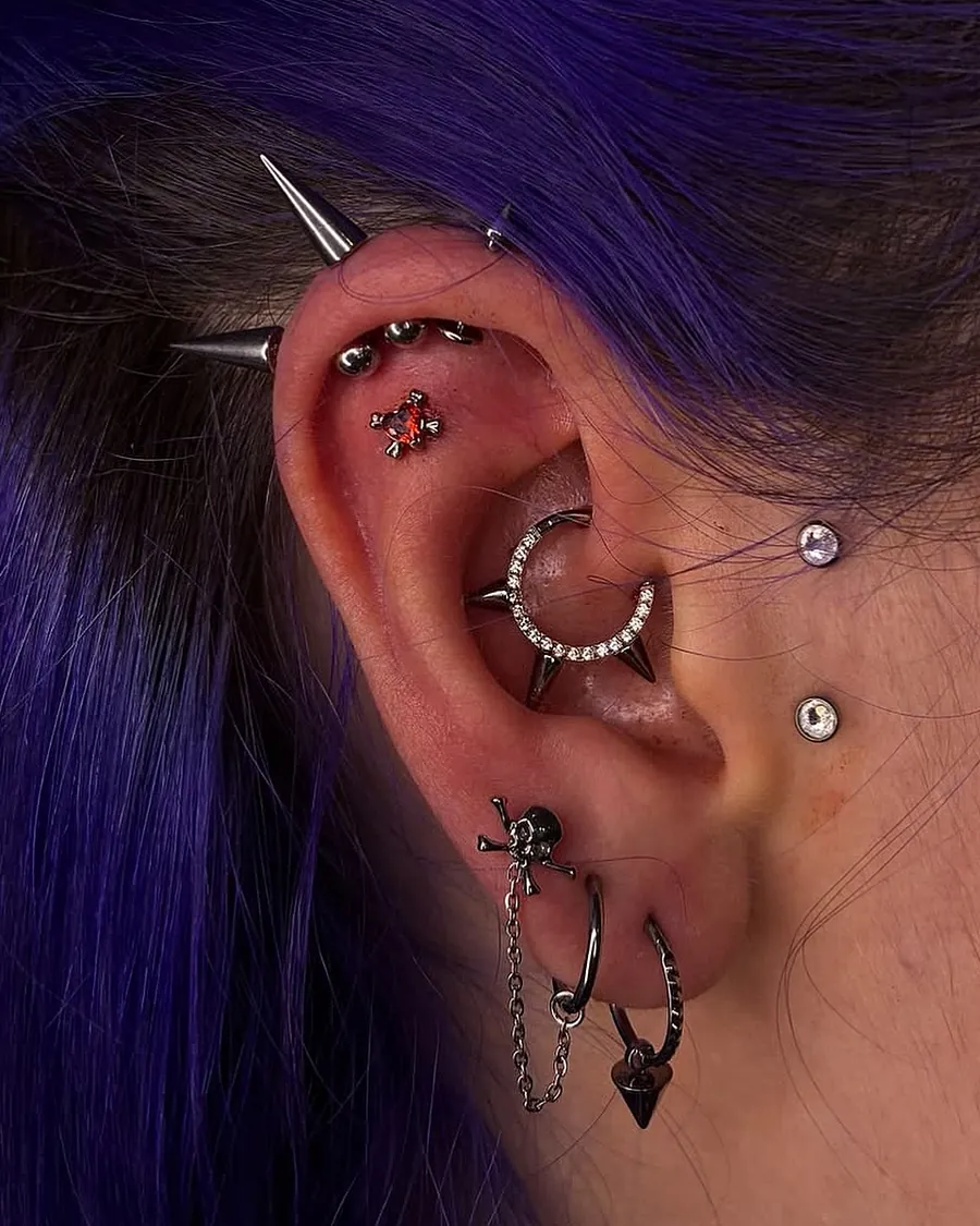 Orelha com curadoria completa: spikes e bolinhas na borda da cartilagem, flat com joia vermelha, daith com argola cravejada, duas joias na frente da orelha e lóbulos com caveira, argolas e corrente